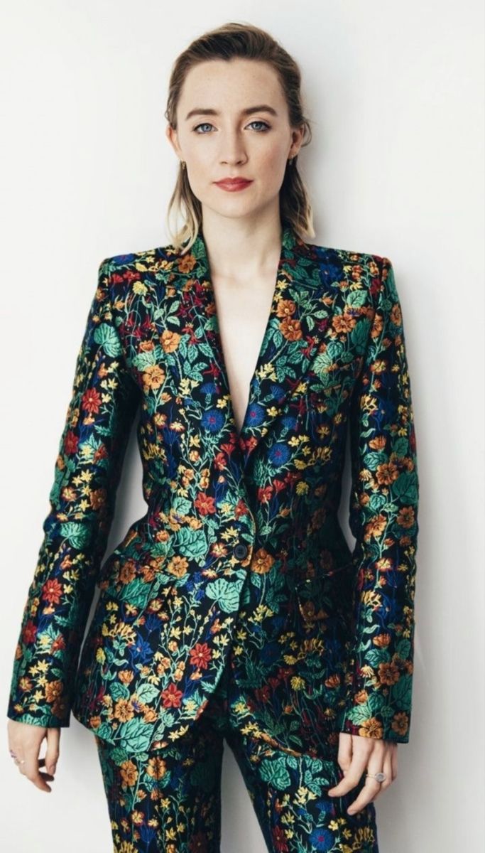a portrait of saoirse ronan wearing a floralpatterned suit against a white background. she is wearing a blazer and matching pants, and has her hair styled back. she is wearing red lipstick and jewelry, and is looking at the camera.