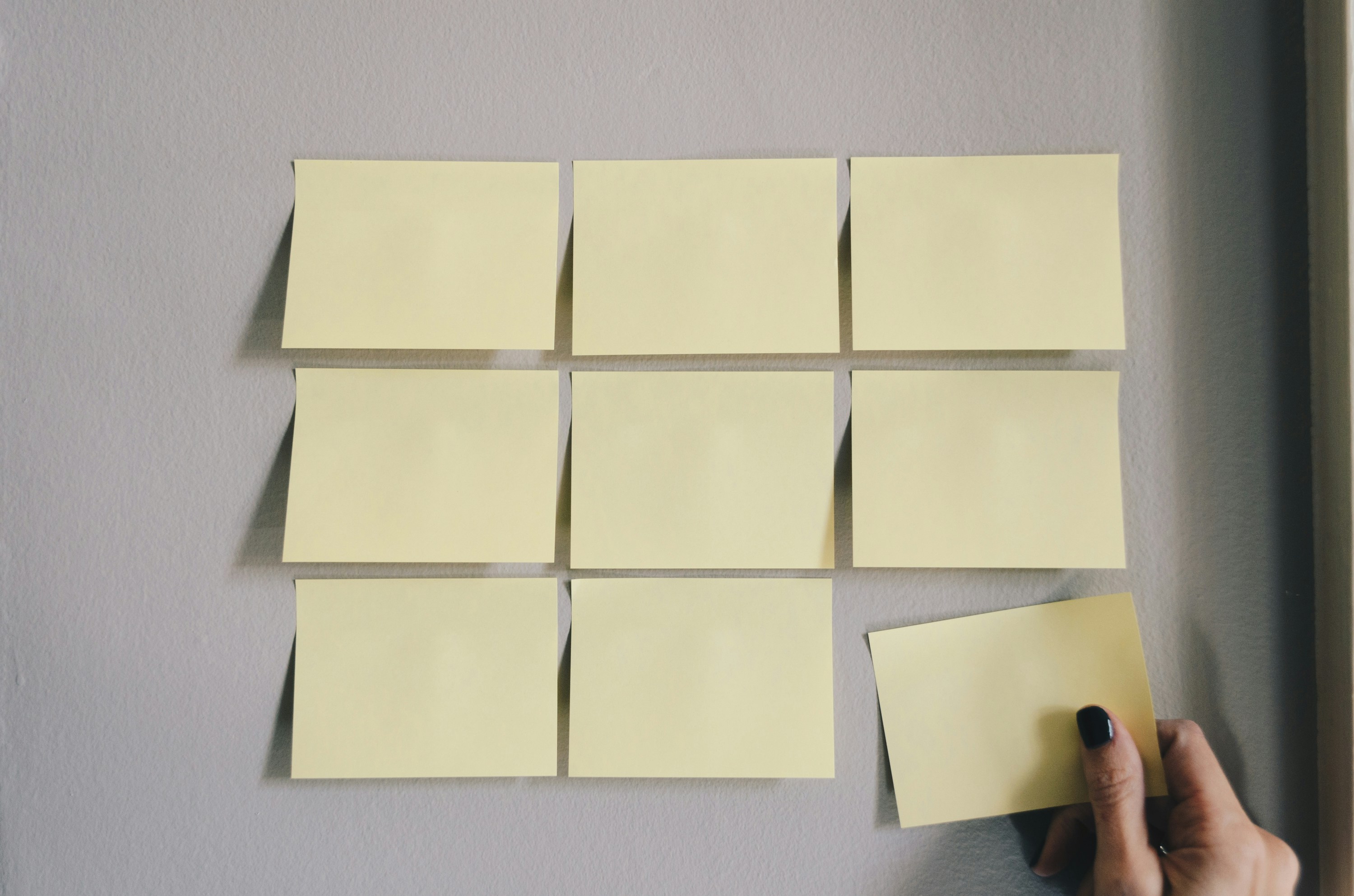 a photo of a gray wall with nine yellow sticky notes arranged in a grid pattern. a person's hand is at the bottom right, holding a tenth sticky note that is partially off the wall.