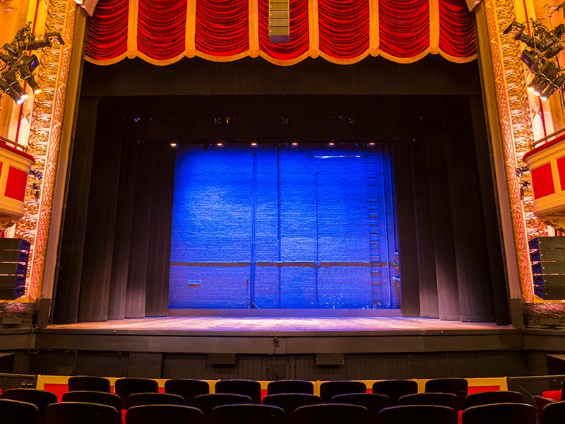 a wideangle shot of a theater stage with dark curtains framing a blue backdrop. red curtains are visible at the top of the stage, and rows of empty seats fill the foreground. the stage lighting illuminates the backdrop and the stage floor.