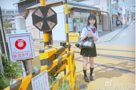 a young woman in a japanese school uniform stands on a sidewalk next to a railroad crossing in an urban environment. the image is taken in broad daylight with natural sunlight illuminating the scene. the perspective is at eye level, providing a clear view of the woman and her surroundings. the overall tone of the image is light and airy.