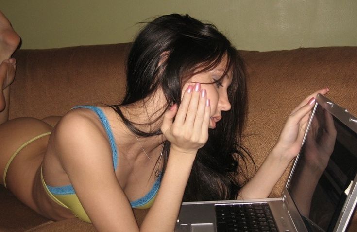 a woman is lying on a brown couch, looking at a laptop. she has dark hair and light skin, and is wearing lingerie. she has pink fingernails. she appears to be indoors.