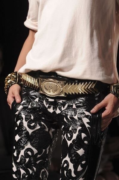 a woman in a white shirt and black pants with a gold belt is posing on a runway.