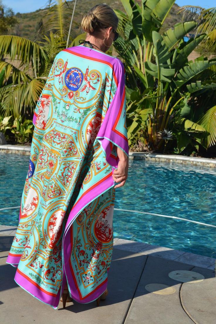 the image shows a woman from behind, wearing a long, flowing silk kaftan. the kaftan is predominantly turquoise with a vibrant baroquestyle print featuring gold, pink, and other colors. the design is intricate and ornate. the woman is standing poolside, near a swimming pool and lush palm trees. she's wearing high heels and appears to be relaxing in a summery or vacation setting.  the overall aesthetic is luxurious and stylish.