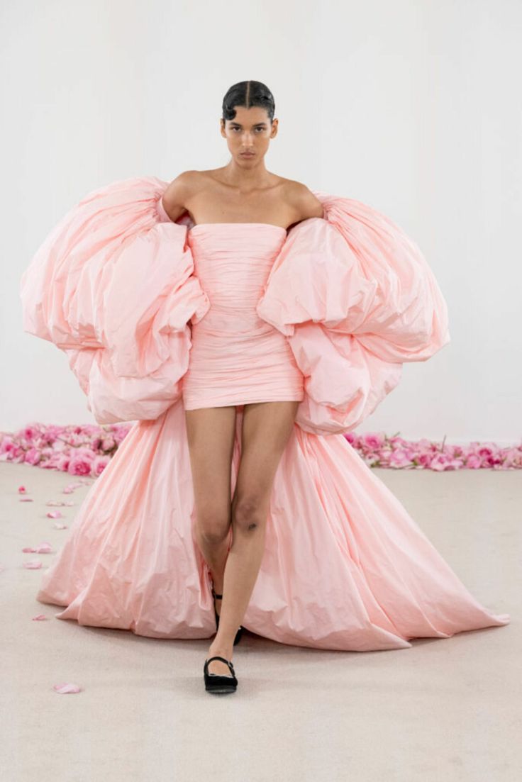 a beautiful woman in a pink, strapless gown poses on a runway. the gown has a large, dramatic puff on each sleeve and the back of the skirt. the gown is very tightfitting on the body, which emphasizes the model's slim figure. she is wearing black, mary janestyle shoes. the runway is covered in rose petals.