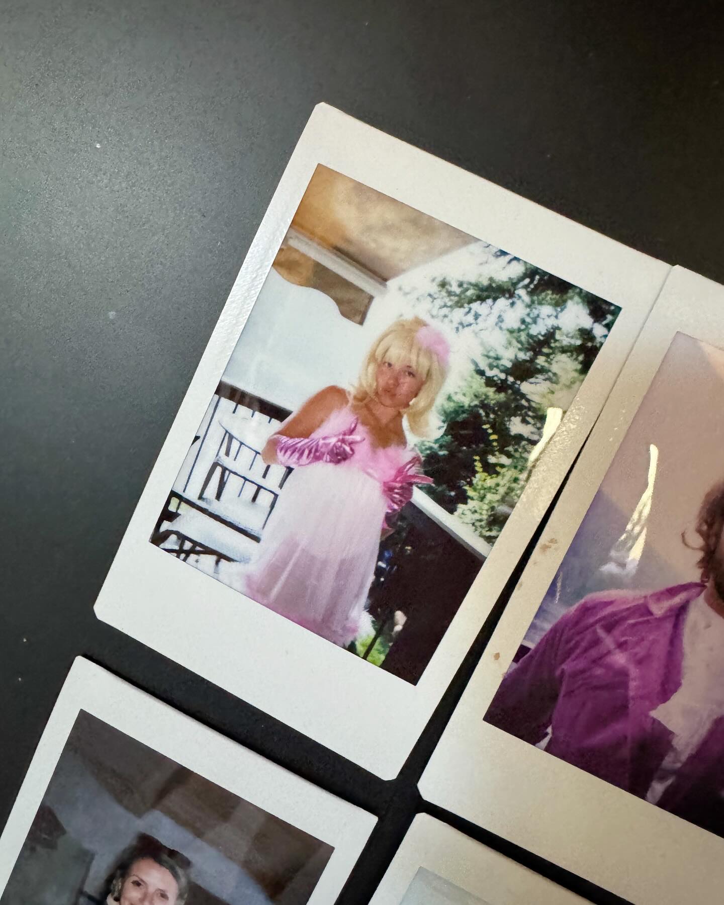 the image shows a collection of instant photos, likely from a photo album, scattered on a black surface. the primary focus is on a snapshot of a woman dressed in a pink outfit, possibly a costume. she is wearing a blonde wig and matching pink gloves, standing outside in front of a railing with trees in the background. the overall aesthetic suggests a retro or vintage style, with the polaroidstyle photos capturing memories of a past event or party. other photos partially visible in the frame show images of other people, adding to the nostalgic feel of the collection.