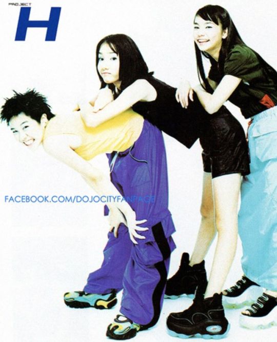 three young asian women are posing together in front of a white background. two of them are smiling and wearing colorful clothing. the woman in the back has her arm around the woman in the middle. all three women are wearing platform shoes. the background says "project h."