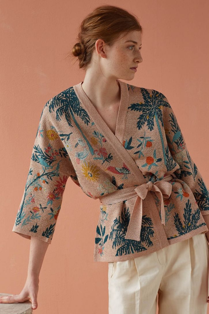 a woman model is showcasing a stylish peachcolored wrap cardigan with a beautiful floral and palm tree print. the cardigan is made of soft knitwear and features a relaxed fit with threequarter sleeves. it's belted at the waist, accentuating the silhouette. the model is wearing offwhite highwaisted pleated pants, completing a chic and casual look. the backdrop is a light pink background that complements the cardigan's colors. the overall aesthetic is soft, feminine, and spring/summer appropriate.