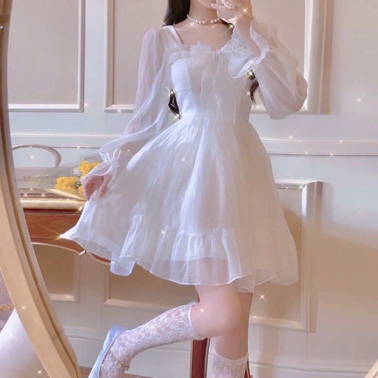 a young woman in a pretty white dress poses in front of a mirror. the dress is aline with long sheer sleeves and a ruffled neckline. the model wears white lace socks and light blue shoes. she also wears a delicate pearl necklace.  the background is an elegant, bright room with classic white walls and molding. the overall aesthetic is sweet, feminine, and romantic.