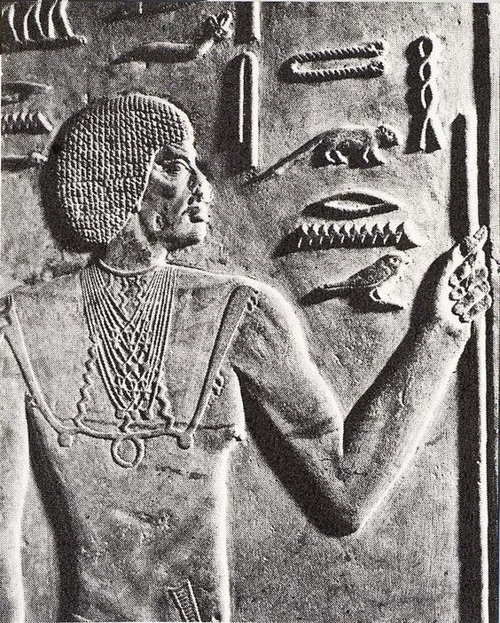 a black and white photo of a carved stone relief sculpture of a man from ancient egypt. the man is shown in profile view, facing to the right, with his right hand raised. he wears a short wig, a necklace, and a kilt. there are hieroglyphs carved to the right of the man.