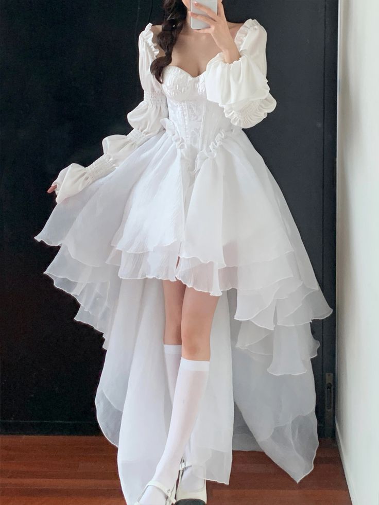 a person is standing indoors taking a selfie, wearing a white dress with long, puffy sleeves and a highlow hem. the dress features ruffles and a layered skirt made of a sheer fabric. the person is also wearing kneehigh white socks and mary jane shoes. the dress has a corseted bodice with decorative details. the overall look is feminine and fashionable.