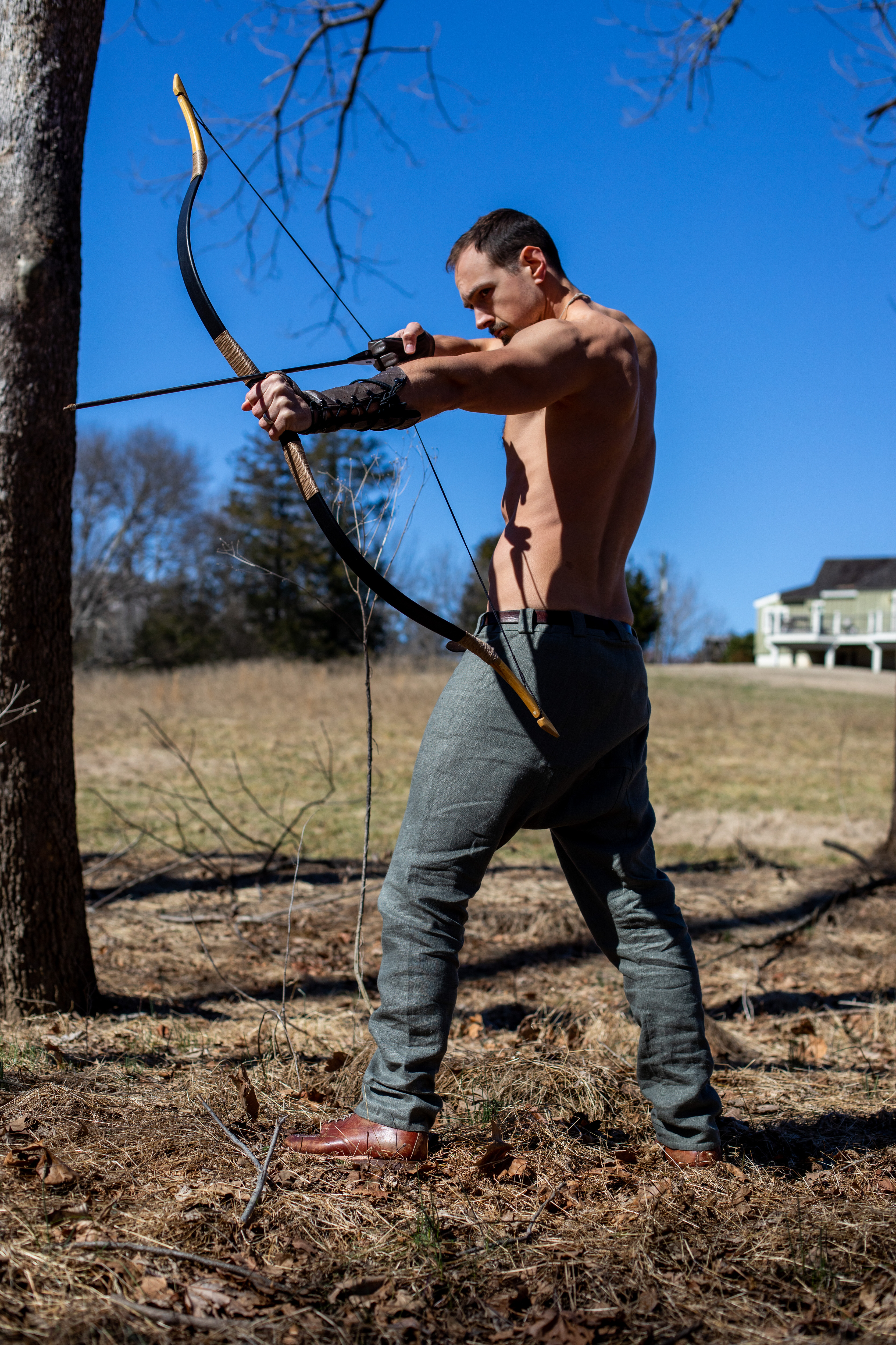 a muscular man with his upper body exposed stands outdoors on a sunny day, aiming a bow and arrow. he is wearing loose gray pants and a leather wrist guard. he's standing in the woods with a house visible in the background.