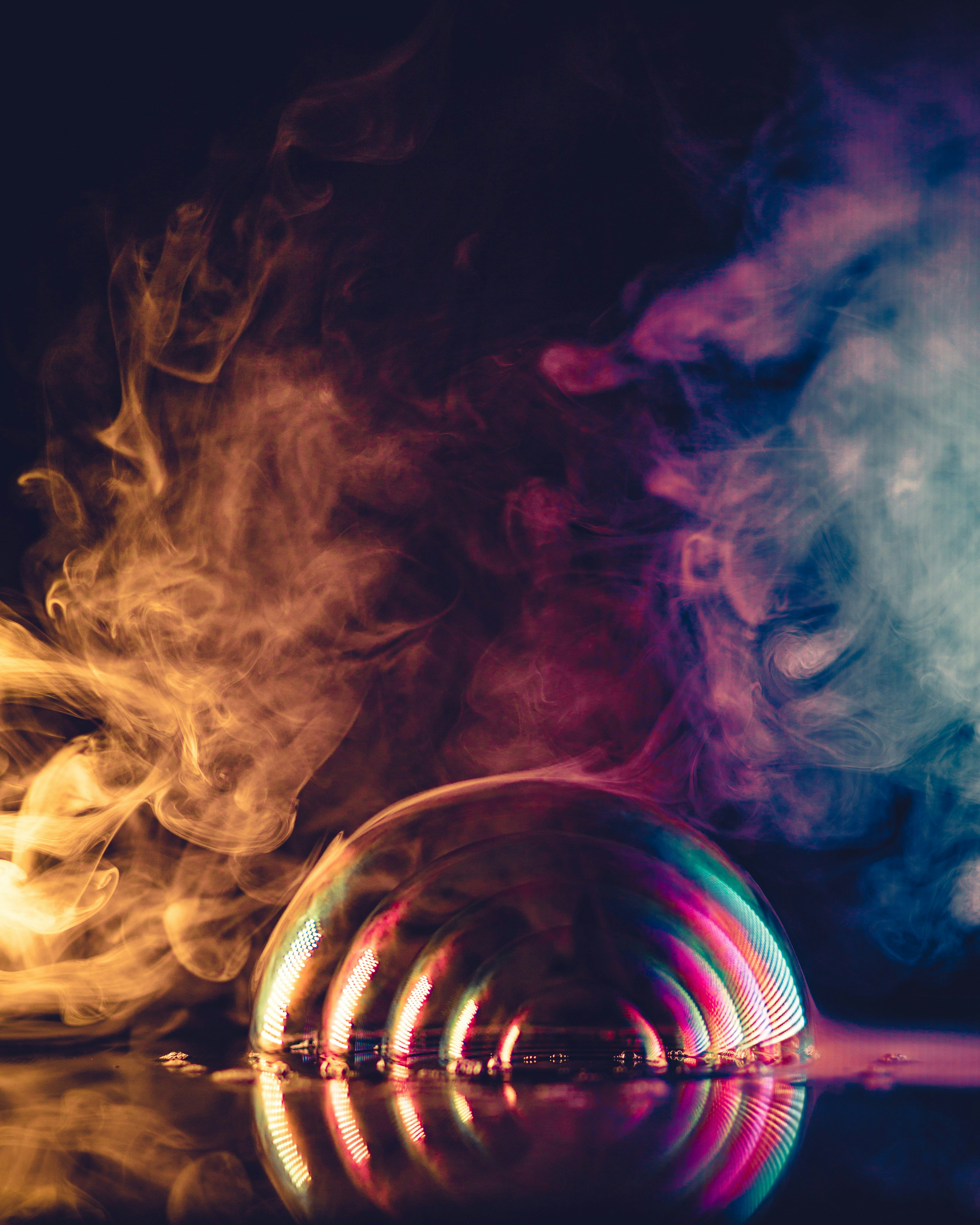 a colorful iridescent bubble sits on a surface with colorful smoke in the background. the image is taken at eye level and the bubble and the smoke is reflected on the surface.