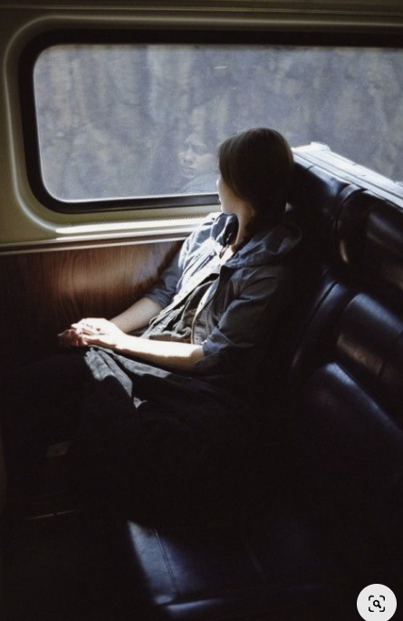 a woman is seated by the window of a train, gazing out at the passing scenery. the natural light illuminates the interior of the train car, casting soft shadows on the woman's face and clothing. she appears lost in thought, perhaps reflecting on her journey.