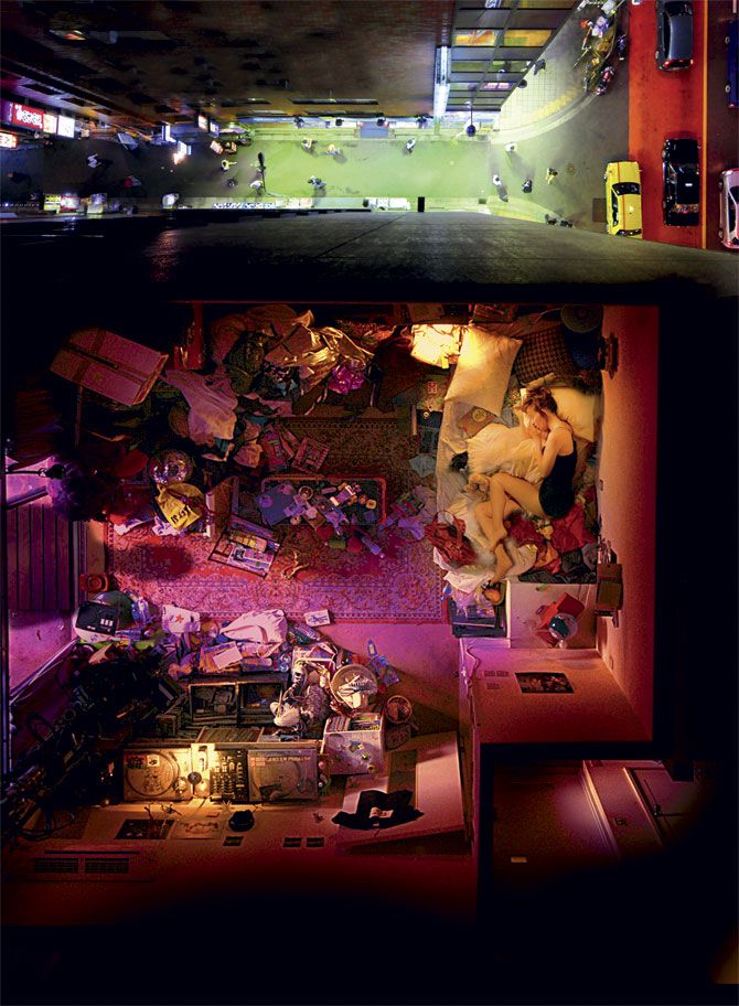 an aerial view of a couple sleeping in a messy bedroom. the room is cluttered with clothes, books, and other items. the city lights are visible through the window. the couple is sleeping in a bed, with the woman's head resting on the man's chest.  the