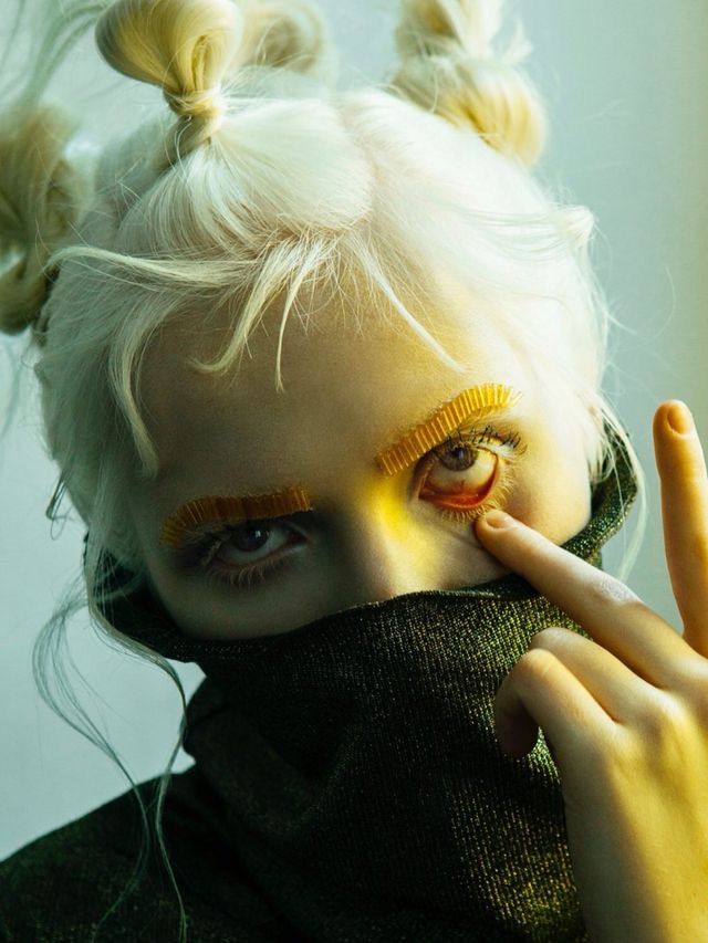 a closeup of a woman with white hair wearing a green turtleneck sweater. she is staring intensely with one eye that has a yellow false eyelash, and the other eye is covered with her hand. her hair is done in two buns.  she is looking fiercely and edgily at the camera, giving off a strong and trendy aura. the green turtleneck sweater is a stylish piece of clothing that adds a touch of sophistication to her look.