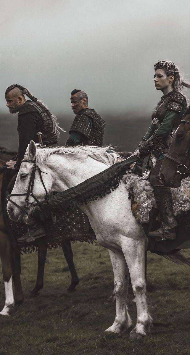 three riders on horseback are on a grassy field under an overcast sky. the riders are dressed in costumes evocative of the viking era, including armor. the horses are white, brown and black. the foreground and background are grass, a hill and fog.