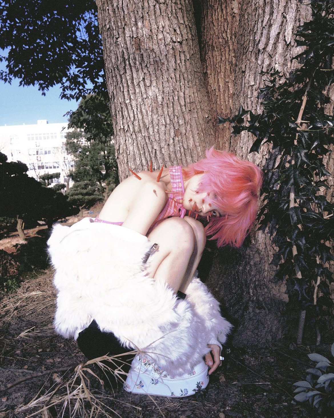 a person with pink hair crouches in front of a large tree. needles stick out of their shoulders and face. they wear a pink and white plaid top, a white fur coat, and floral platform shoes. the background shows a building and some greenery.