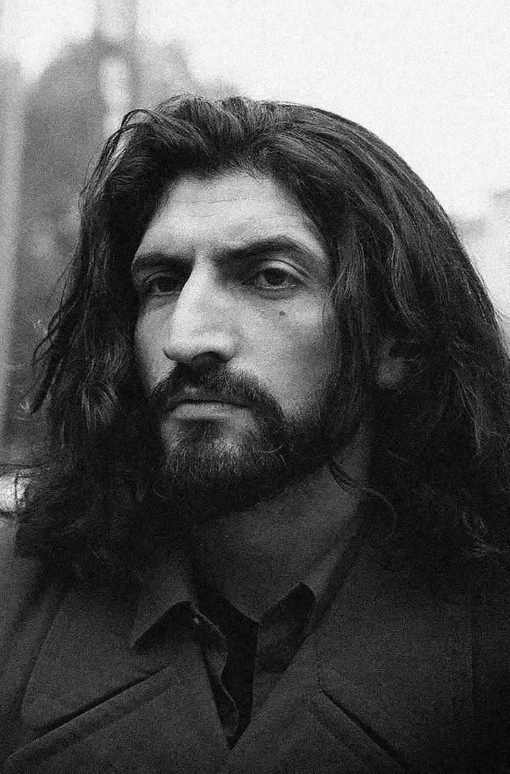 a black and white portrait of a man with long dark hair and a beard, looking directly at the camera with a serious expression. he is wearing a collared shirt and a coat.