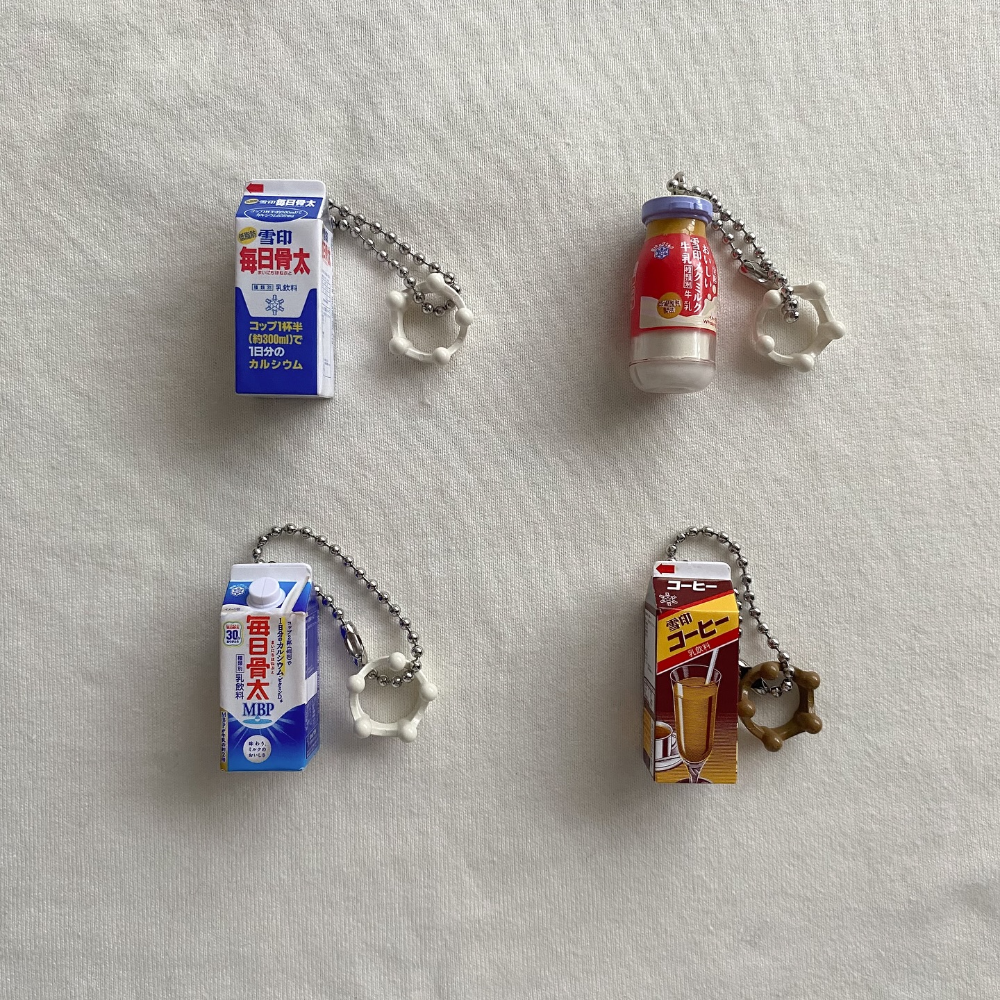 the image shows four different milk/coffee keychain trinkets laid on a white surface. the trinkets are different sizes and shapes, but all have a similar design. each keychain is attached to a small ball chain and has a white or brown ring attached to the end. the keychain trinkets are all made to look like small cartons or bottles of milk or coffee. the cartons and bottles are decorated with japanese writing and images.