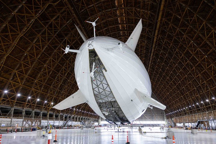 a large white airship, or dirigible, is suspended inside a hangar. the airship is secured with cables and ropes. it is positioned vertically, with its nose pointed upwards. the hangar is large and has a wooden truss roof. the floor is concrete and there are workers in the distance.  the airship is a modern piece of engineering, and the hangar is a testament to the size and scale of the aircraft. this photo captures a moment of preparation for a future flight.