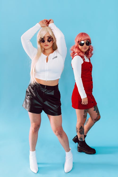 two young women are posing against a bright blue background.  one woman is wearing a white cropped zipup shirt, black leather shorts, and white ankle boots. she has blonde hair and is wearing decorative sunglasses. the other woman is wearing a red mini dress, white longsleeved shirt underneath, and black chunky boots. she has pink hair and is also wearing sunglasses.  both women appear confident and stylish. the overall aesthetic suggests a 90sinspired fashion look.