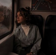 a young woman with curly brown hair sits on a train, gazing thoughtfully out the window.  the lighting is dim and atmospheric, casting a melancholic mood over the scene.  she is wearing a blue shirt and her expression suggests a sense of contemplation or introspection.  the color palette is muted and earthy, with shades of blue, gray, and brown dominating the image.