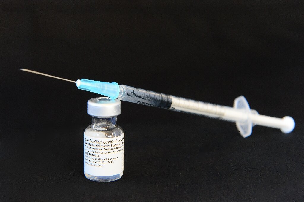 the image shows a vial of pfizerbiontech covid19 vaccine with a syringe placed on top of it against a dark background. the syringe has a blue cap. the label of the vial is visible, with text mentioning dilution, storage temperature, and emergency use authorization.