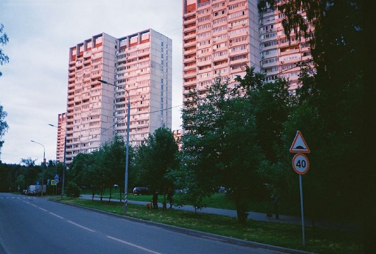 the image depicts a quiet residential street in what appears to be a russian or eastern european city. two large, multistory apartment buildings, characteristic of sovietera architecture, dominate the background. the buildings are lightcolored, appearing pinkish in the evening light.  lush green trees line the street, which is relatively empty except for a few vehicles in the distance. a speed limit sign indicating 40 km/h and a hump warning sign are visible on the righthand side of the road. the overall atmosphere is calm and peaceful, capturing the quiet ambiance of a residential neighborhood at dusk. the image has a grainy, slightly vintage quality suggestive of film photography.