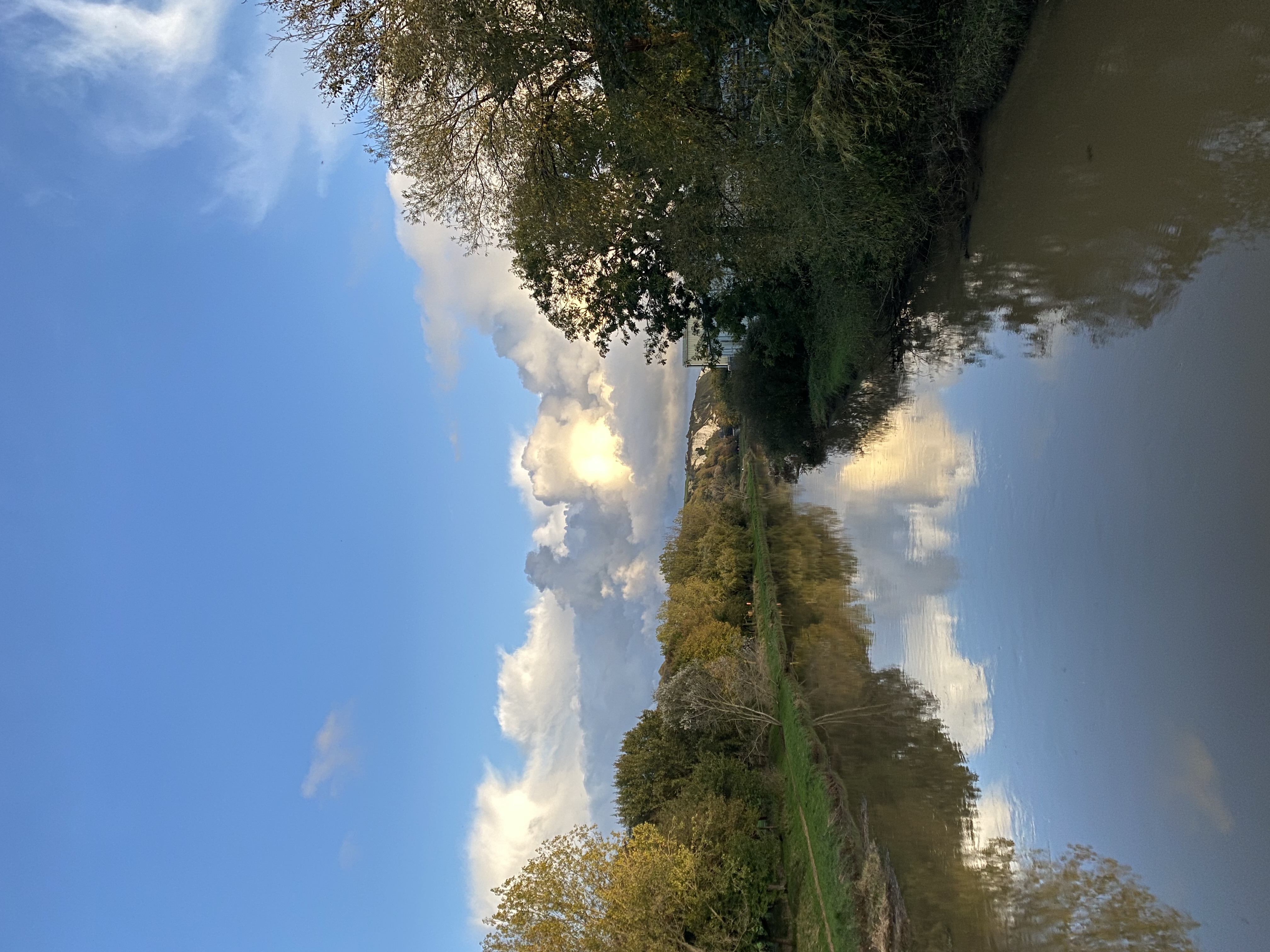 a tranquil river scene with a blue sky and white fluffy clouds reflecting in the still water.  the river flows gently towards the horizon with green trees lining its banks on both sides. the sky is bright blue with patches of white clouds casting shadows on the water below. the scene is peaceful and serene, with a soft golden light illuminating the landscape.