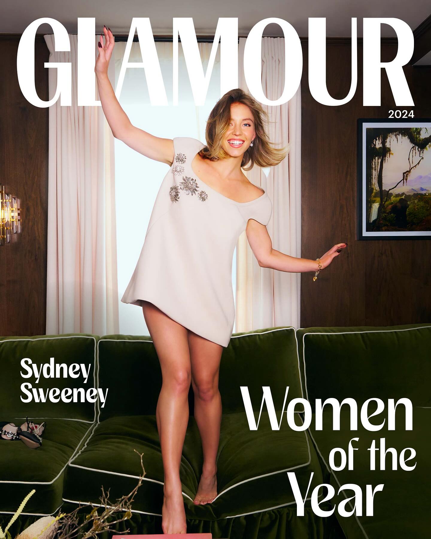 the image is a glamour magazine cover featuring actress sydney sweeney as their "women of the year".  sweeney is playfully perched on a dark green velvet sofa, wearing a light beige mini dress. she's smiling and looking directly at the viewer. the background features a portion of a room with curtains and an art piece on the wall. the magazine's title, "glamour," and the year "2024" are prominently displayed. the text "sydney sweeney" and "women of the year" are also visible.