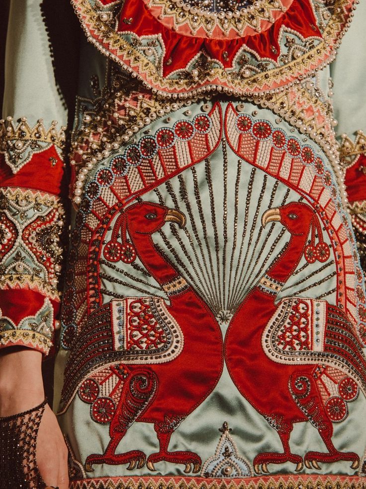 closeup shot showcases a richly embellished dress, possibly haute couture.  the dress features a light teal base fabric adorned with intricately beaded peacocks in shades of deep red and gold. the peacocks are centrally positioned, surrounded by elaborate radiating patterns of beads and embroidery. the neckline and cuffs show additional layers of ornate embellishments and beadwork. the overall effect is one of extreme luxury and intricate craftsmanship, hinting at a highfashion or possibly even historical context. a portion of a model's hand and arm is visible at the bottom left, adding a sense of scale to the garment's detail.