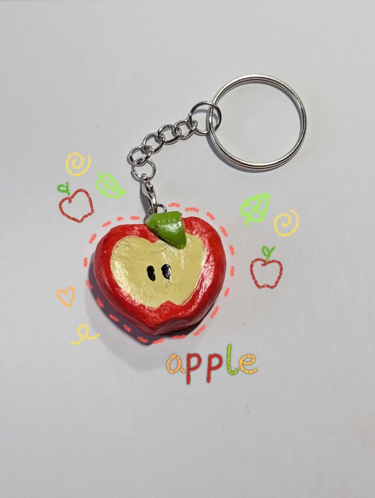 a handmade keychain of an apple made from clay. the apple is red with a yellow core and a green leaf. it is attached to a silver keychain. the image is decorated with doodles of apples, leaves, swirls, a heart, and the word "apple."
