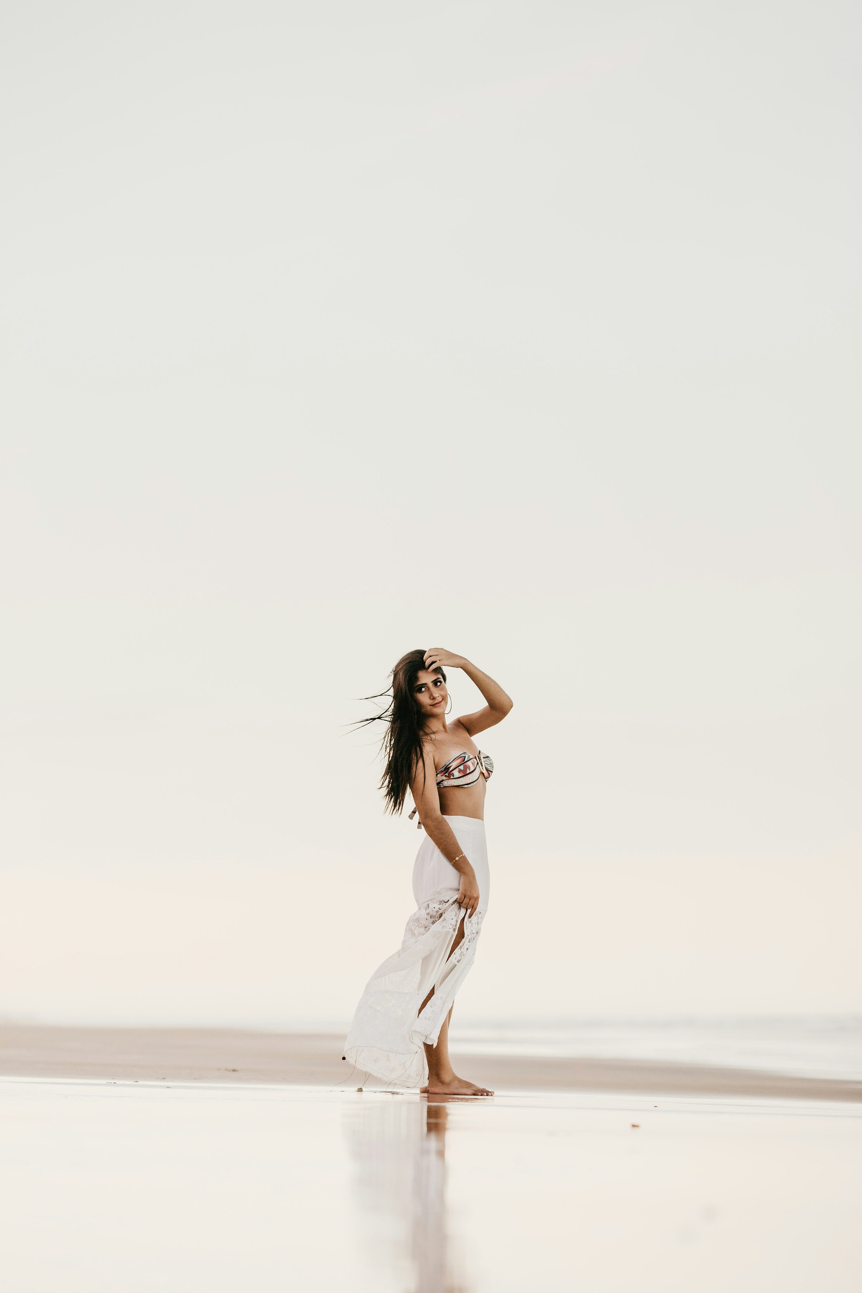 a full shot of a woman on a beach, standing in the shallow water. she's wearing a white skirt and a patterned bikini top. her long, dark hair is loose. the sky and ocean are a pale, clear color. the overall feeling is one of peace and serenity.