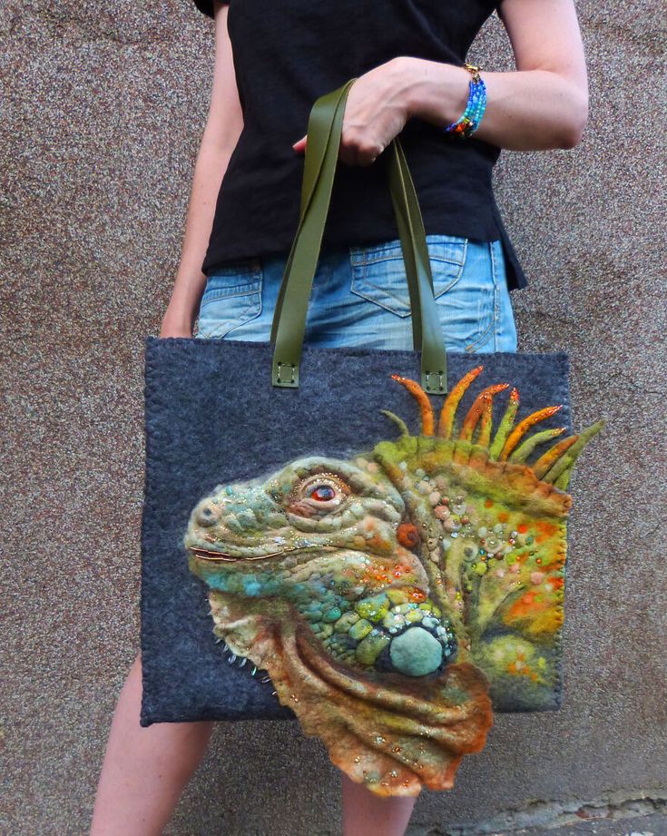 a woman is holding a unique tote bag with a handfelted iguana on the front. the bag is made of grey felt and has green leather handles. the iguana is a vibrant mix of green, orange, yellow and blue, with a textured surface. the bag is a perfect accessory for any fashionconscious individual who appreciates handmade art and unique pieces.