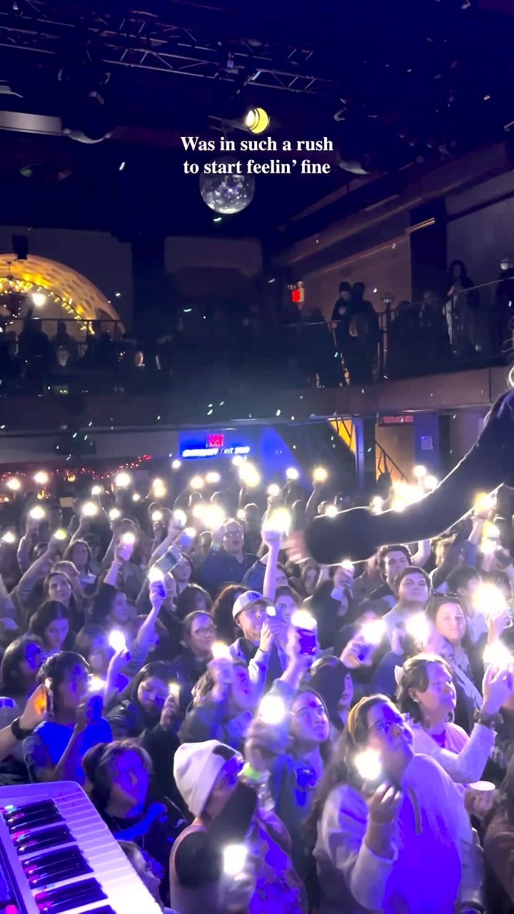 a crowd of people at a concert are holding up their cell phones with the flashlights on. there is a disco ball hanging from the ceiling and a keyboard in the foreground.