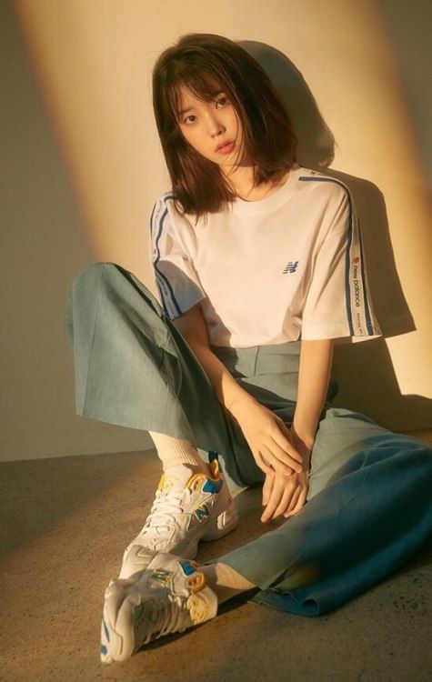 a woman is sitting on the floor with her legs crossed. she's wearing a white shirt with blue trim on the sleeves and blue pants. she's wearing a pair of white, yellow, and blue new balance sneakers.  she has straight, dark brown hair with a center part and bangs. she's looking at the camera with a soft expression on her face.  the lighting is natural and the background is a light brown wall.