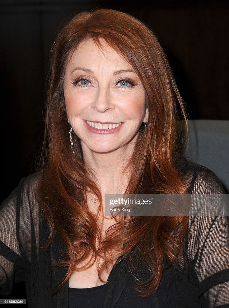 a closeup headshot of a woman with long red hair, smiling. she's wearing a black dress and has a necklace on.  the background is blurred and dark.