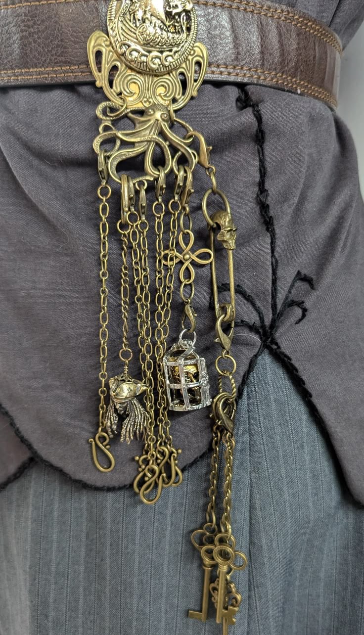 a closeup shot shows a steampunkstyle belt with various charms and chains. a large, ornate brass belt buckle features an octopus design. attached to the buckle are several chains, each holding different charms, including a cage, a skull, and a set of keys. a brooch or pin with a skull design is also visible. the belt is worn over a gray garment with a decorative black stitch detail.