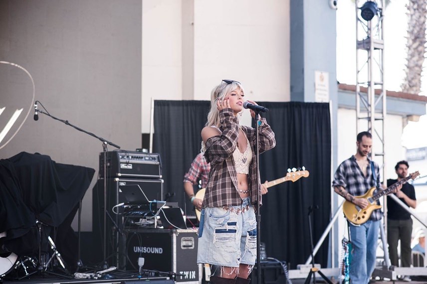 a woman sings into a microphone on a stage with a band behind her. she is wearing a plaid shirt, denim shorts, sunglasses, and a crocheted top. the band includes a guitarist and a bassist. there are amplifiers and other equipment on the stage.
