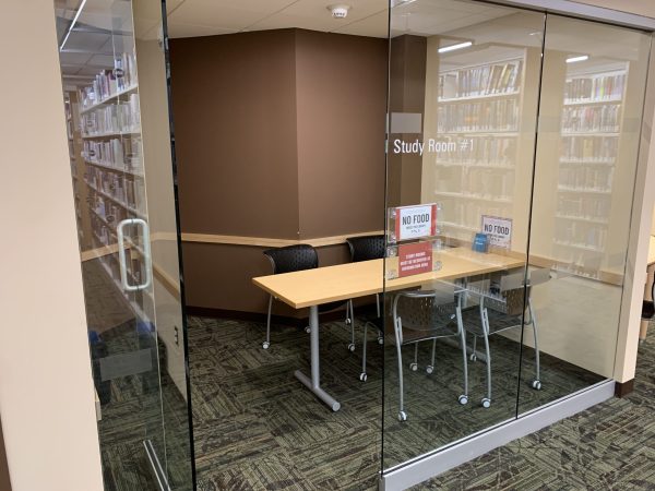 a small study room in a library, with a table, four chairs, and bookshelves on the walls. there are two signs that say "no food" and a sign that says "study room #1" on the glass door. the study room is separated from the rest of the library by glass walls. the floor is carpeted in a brown and green pattern. there is a door to the left of the study room that leads to the rest of the library.