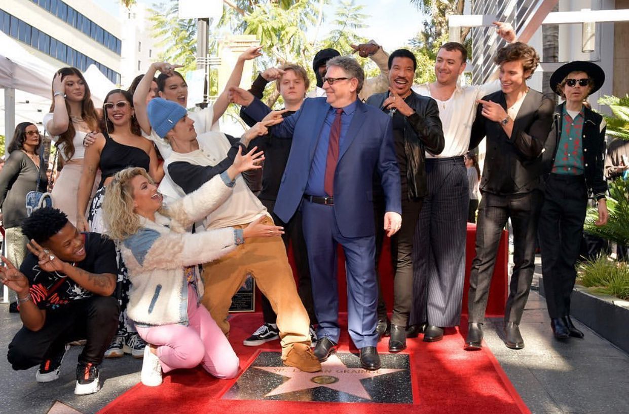 a group of people are gathered on the hollywood walk of fame around the star of sir lucian grainge. several individuals are pointing at the star while posing for a photo. there are men and women dressed in both formal and casual attire, including a person in a blue suit and others in more relaxed outfits. a red carpet is spread out around the star.