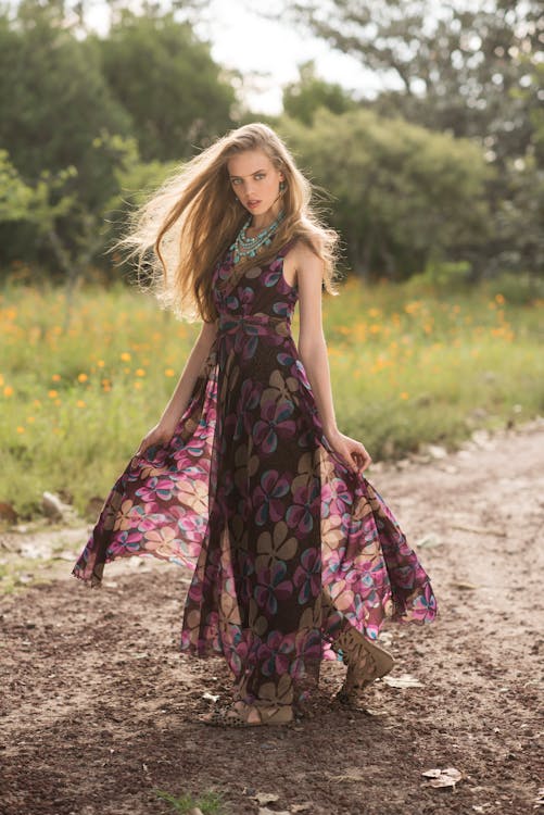 a blonde woman is standing on a dirt path in a field, wearing a brown floral print maxi dress, turquoise necklace and sandals. she is holding the edges of her dress, allowing it to billow slightly. the field is dotted with small yellow flowers and the background consists of green trees and foliage, bathed in sunlight. the woman's long hair is flowing in the breeze.