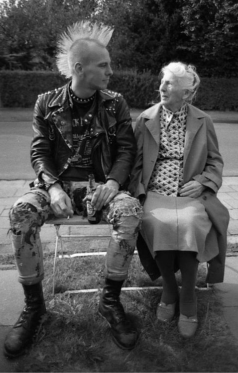 a black and white photo of a young punk rocker with a mohawk sitting on a bench with a older woman, possibly his grandmother. they appear to be chatting and enjoying each other's company. the photo captures a classic generational gap between the two.