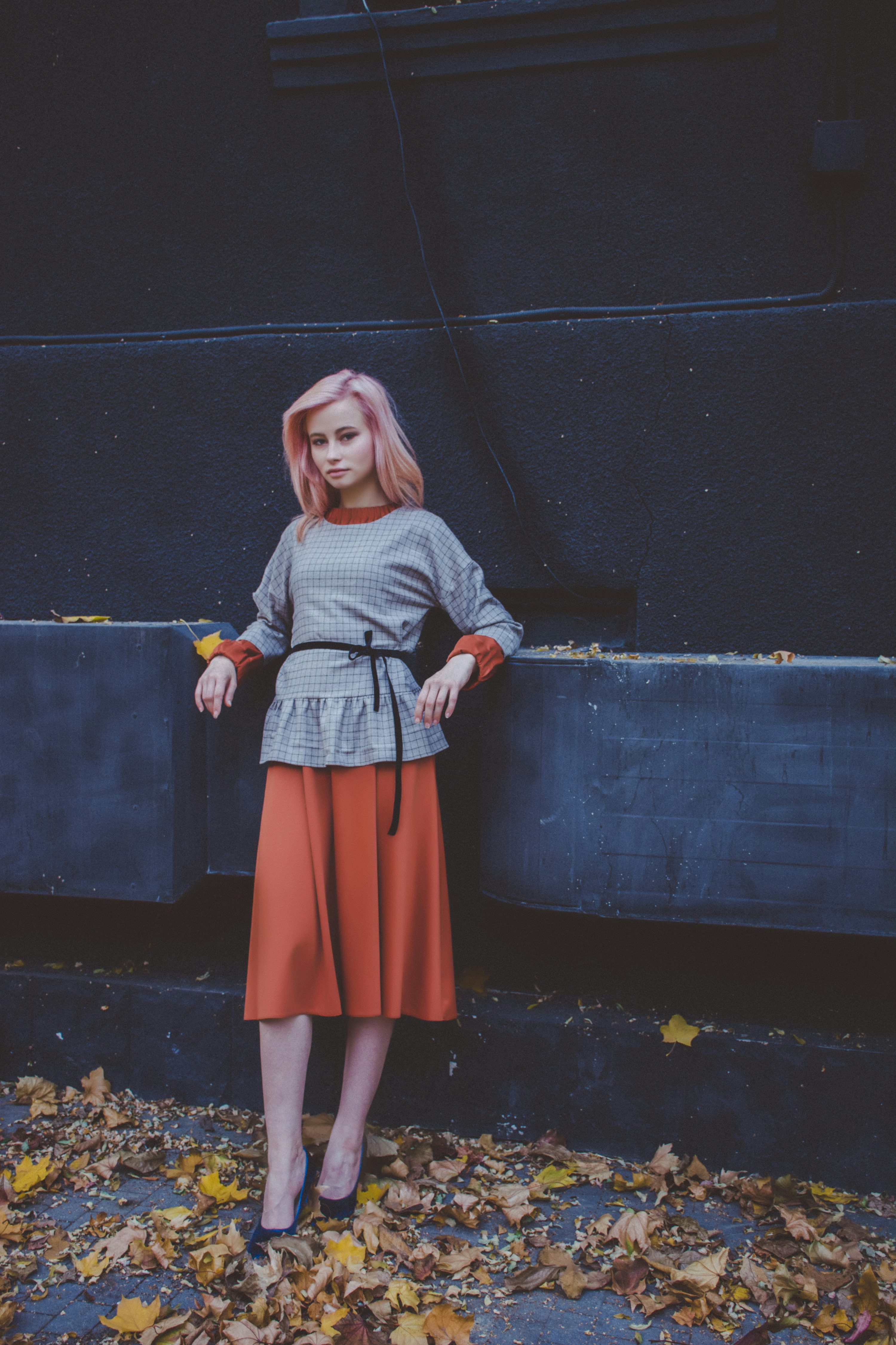 a young woman with pink hair stands against a dark wall, leaning slightly against it.  she's wearing a grey checkered sweater, a rustcolored midi skirt, and black heels.  fallen autumn leaves are scattered on the ground around her feet. the overall mood is stylish and autumnal.