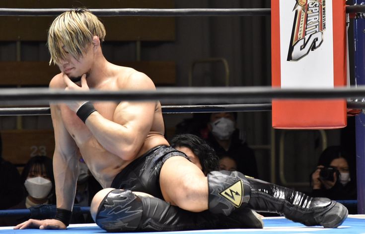 male wrestler, with blond hair, sits in a wrestling ring, his hand to his chin in a thoughtful pose. he's wearing black wrestling shorts and boots, and his muscular physique is visible. another wrestler's leg is draped over his own. spectators, some wearing masks, are visible in the background. the image is taken in an indoor arena, with the branding of a wrestling company visible.