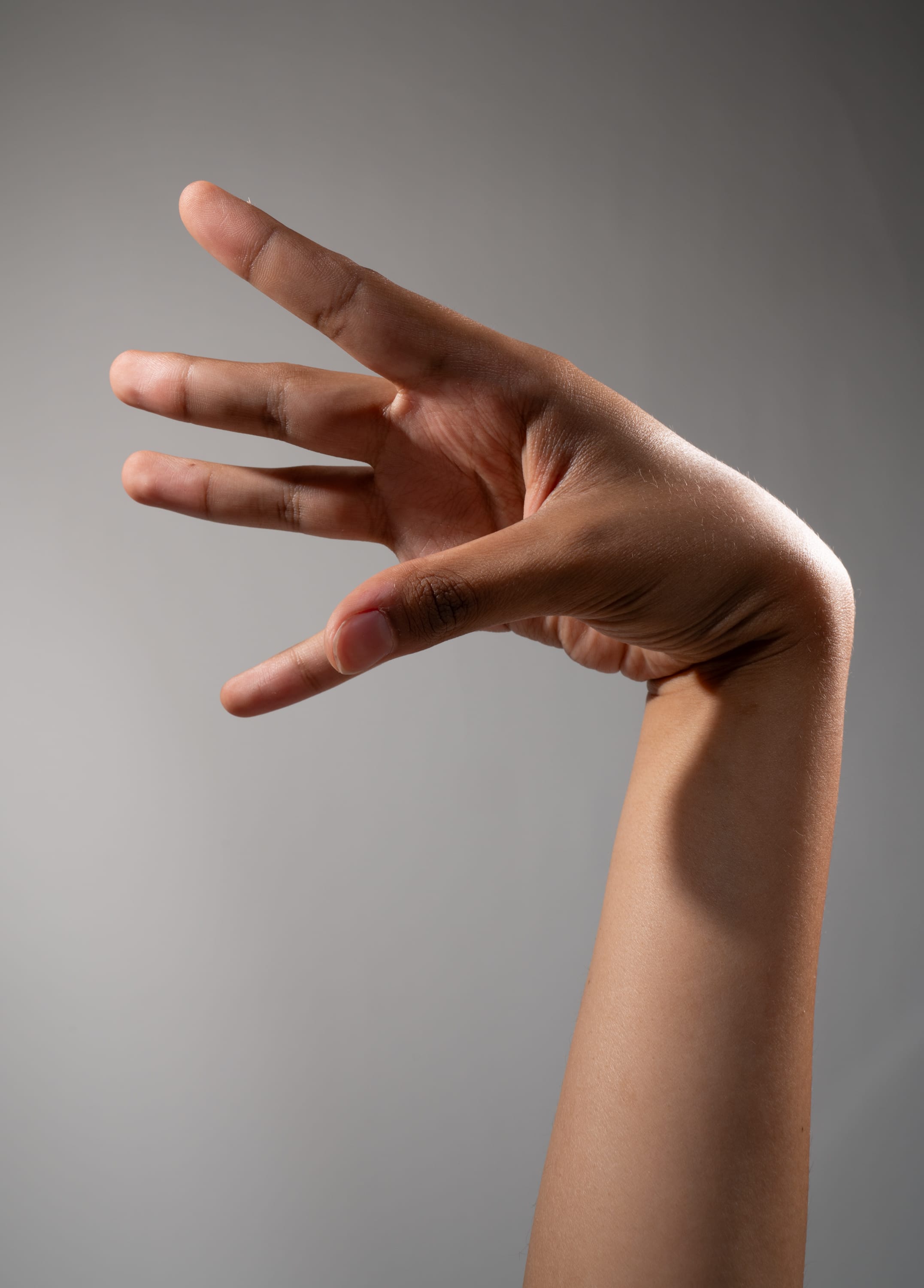 a hand with dark brown skin is extended towards the viewer, palm facing out.  the fingers are extended. the background is a solid light gray, and the arm is illuminated by studio lighting.