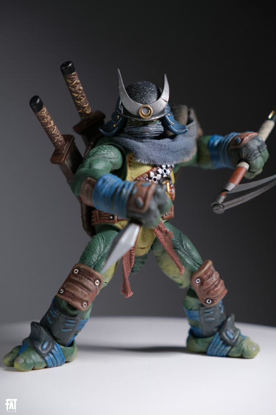 a teenage mutant ninja turtle action figure dressed as a samurai. the figure is wearing a helmet and has swords strapped to its back. it is holding a sai in one hand and a knife in the other. it is wearing gear on its arms and legs.