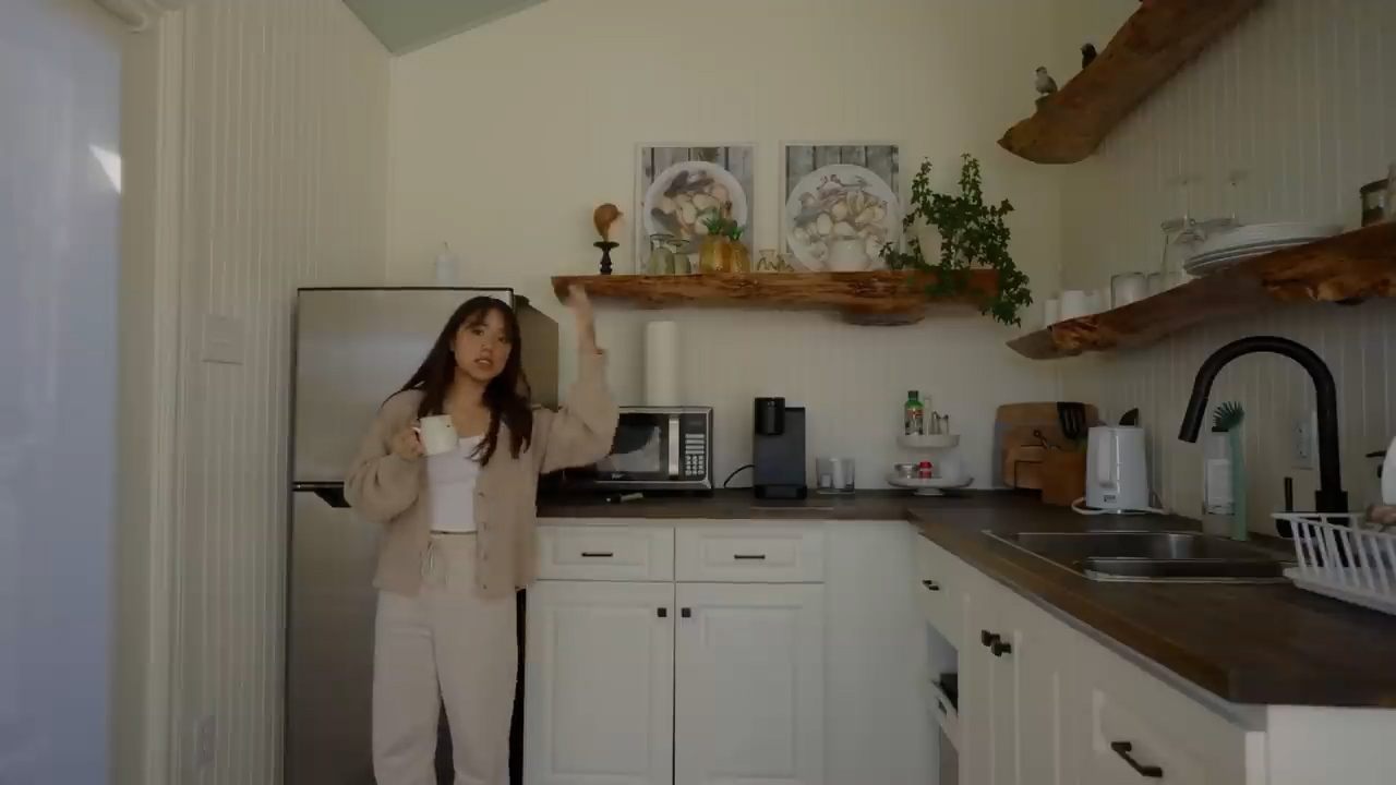 a woman stands in a bright, white kitchen with rustic wood accents. she is holding a mug and appears to be explaining something. the kitchen features white cabinets, a stainless steel refrigerator, and open shelving with decorative items. the countertops are a dark wood and there is a window on the far left side of the image allowing natural light to brighten the space.