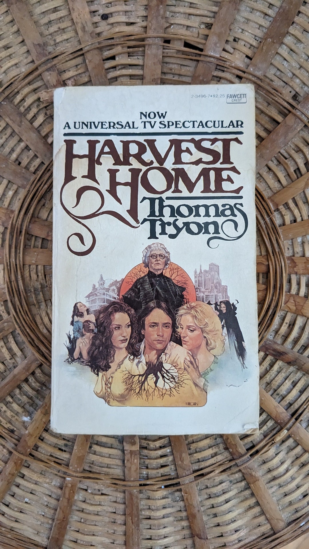 a closeup shot of a vintage paperback book titled "harvest home" by thomas tryon.  the book cover features an illustration depicting a family, possibly in a historical setting, with a large tree at its center. the cover also notes that it's a "universal tv spectacular," suggesting it was adapted into a television miniseries. the book rests on a wicker basket, which provides a textured background. the overall aesthetic suggests the book is from the 1970s.