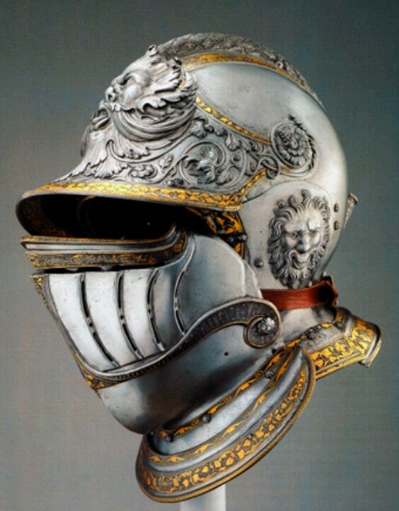 a closeup view of an ornate silver and gold helmet, likely from the renaissance era. it features intricate engravings, lion head decorations, and a movable visor. the helmet is displayed on a stand against a neutral gray background.